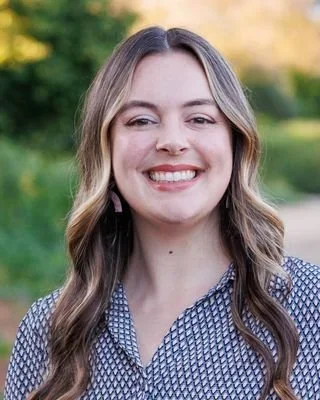 A young woman with long, wavy brown hair smiling outdoors in a park with trees and sunlight in the background.