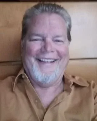 A smiling man with light-colored hair and a beard, wearing a tan shirt, sitting in front of a wooden background.