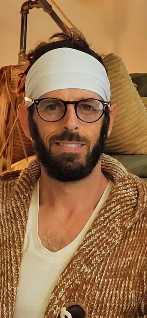 A man with glasses, a white headband, and a beard smiling, wearing a white shirt and a brown knitted cardigan, sitting on a couch with pillows in the background.
