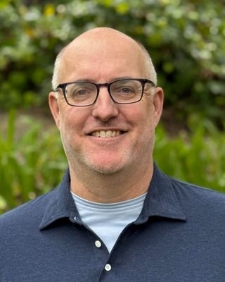 A man with glasses smiling outdoors with greenery in the background.