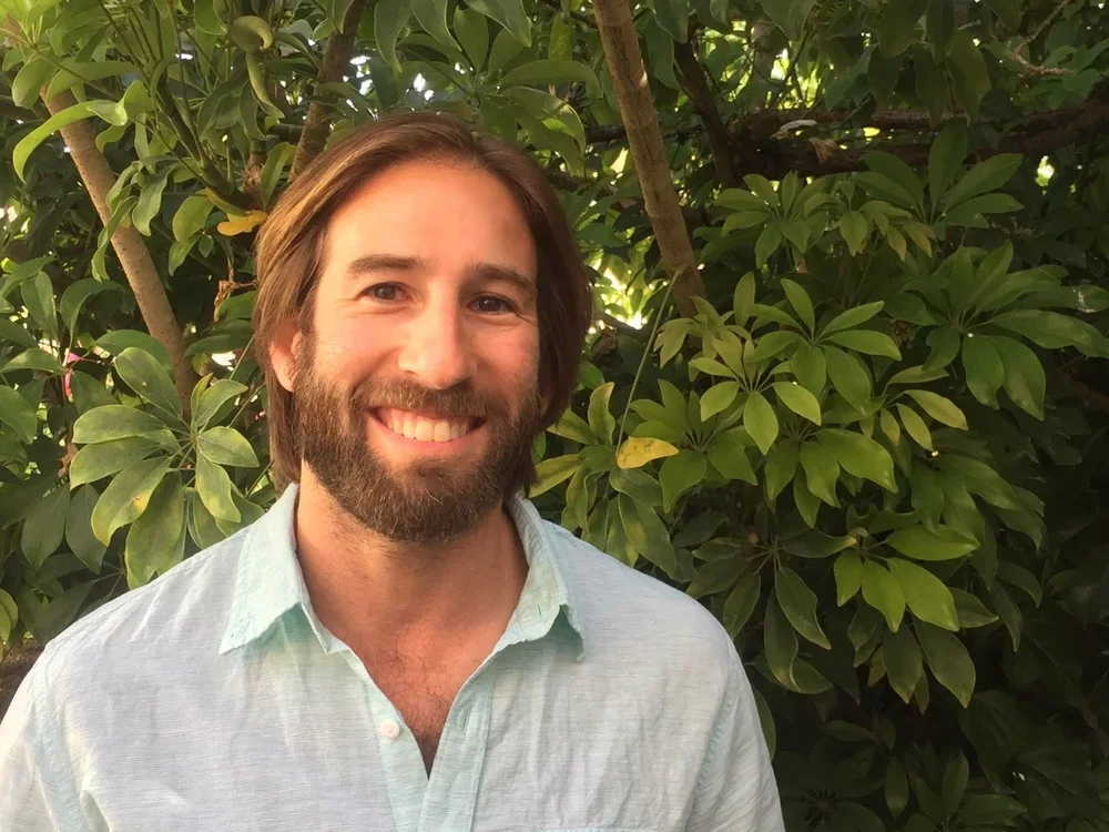 A smiling man with a beard and brown hair, wearing a light blue button-up shirt, standing outdoors in front of lush green foliage.