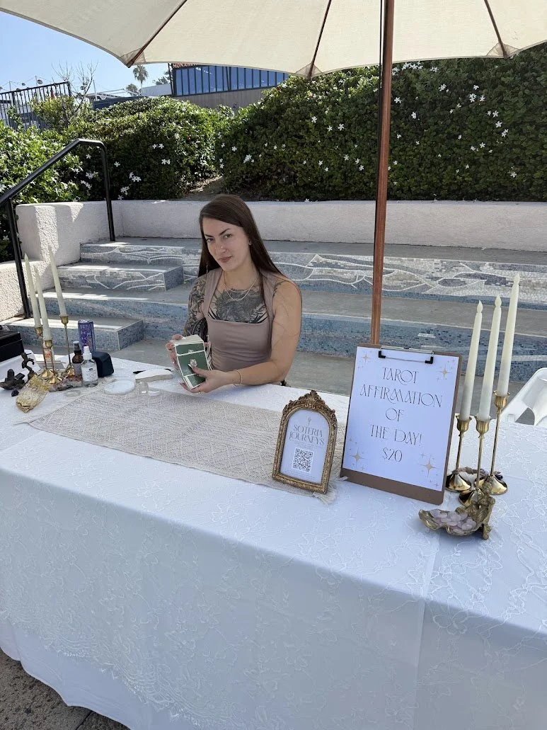 A woman sitting at an outdoor table with a white tablecloth, tarot cards in her hands, under a large umbrella. The table has candles, bottles, and a framed sign that reads 'Tarot Affirmation of the Day! $20' and a decorative frame with a QR code.