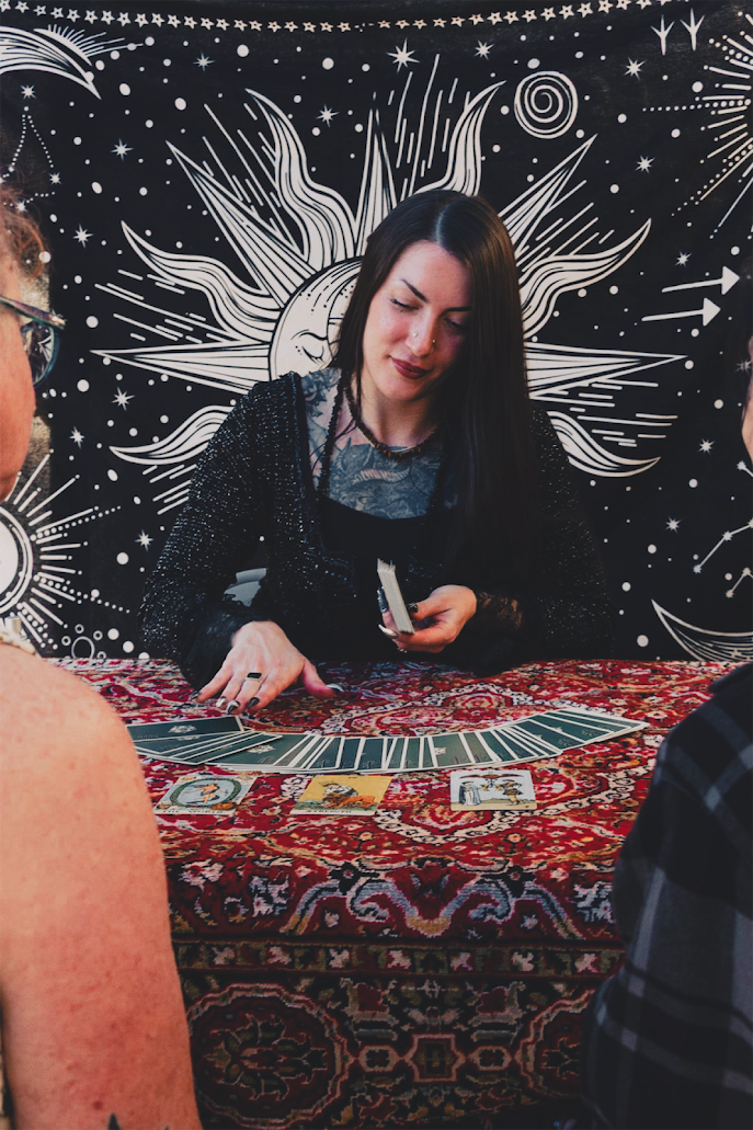 A woman with long dark hair and a black lace top participates in a tarot card reading session, seated at a decorated table with tarot cards and oracle cards, with an artistic black and white celestial-themed tapestry in the background.