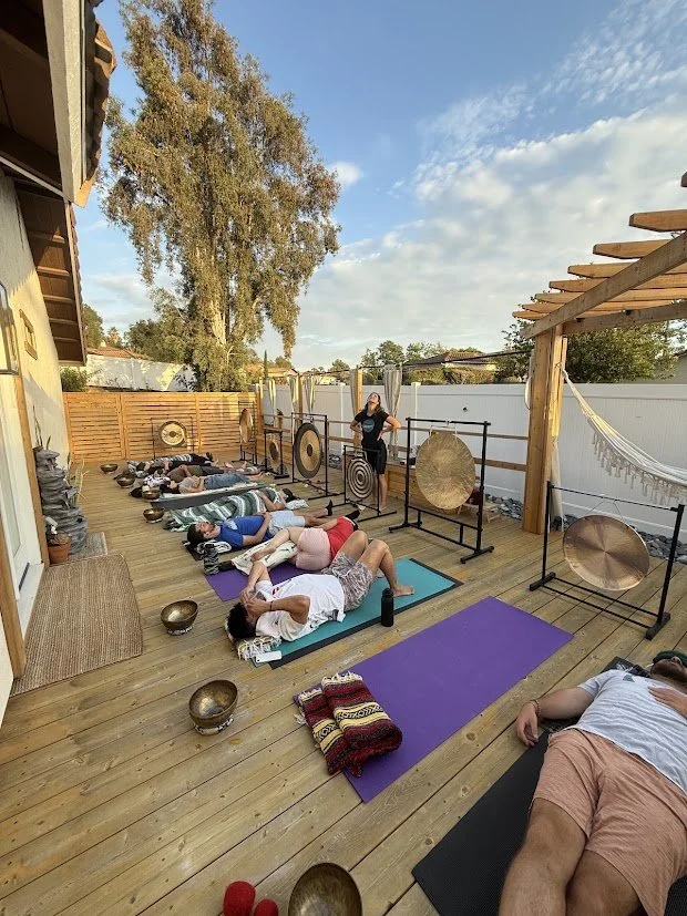 People participating in a sound healing or meditation session on a wooden deck outdoors, with gongs, Tibetan bowls, yoga mats, and a hammock, under a partly cloudy sky.