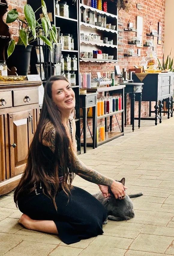 A woman with long brown hair and tattoos on her arm, sitting on a carpeted floor, smiling and petting a gray cat, in a vintage-style room with brick walls, wooden furniture, and shelves with jars with colorful and mystical items.
