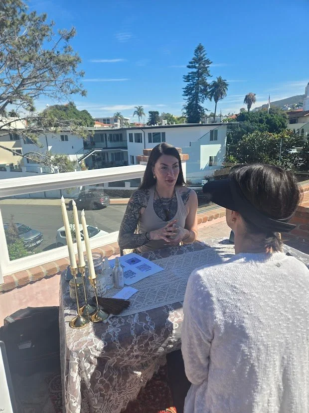 Two women sitting at a table on a balcony, engaged in a conversation. One woman has dark hair, tattoos on her arms, and is wearing a sleeveless top. The other woman has her hair covered with a large black headwrap and is wearing a white sweater. The table has a lace tablecloth, candles, and papers. In the background, there are trees, houses, and a clear blue sky.