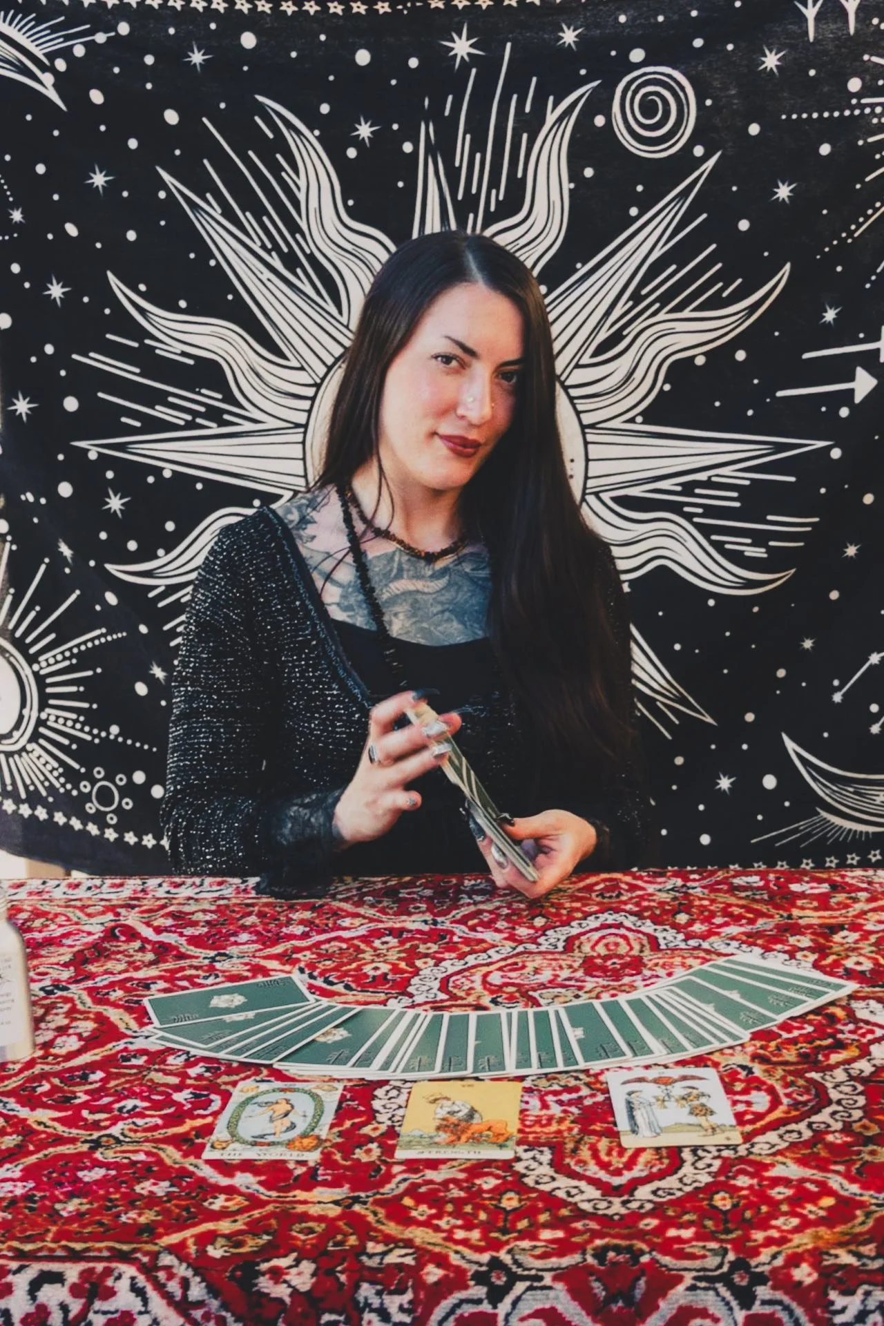 A woman with long dark hair and tattoos is sitting at a table covered with a red ornate cloth, holding tarot cards. Behind her is a black wall with white celestial-themed artwork, including stars, spirals, and abstract lines.