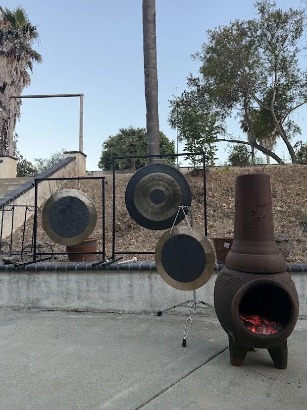 Three gongs of different sizes on stands and a clay chimenea outdoor heater with a fire burning inside, outdoors with trees and a clear sky in the background.