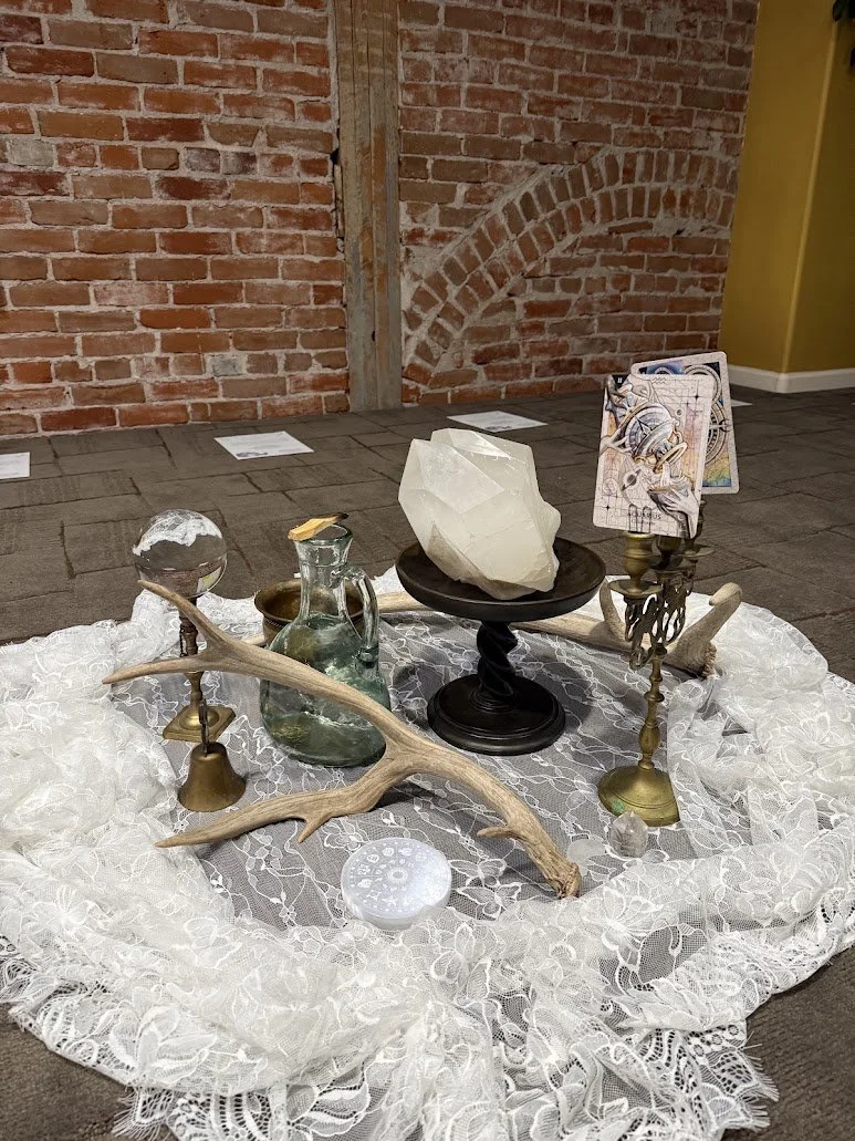 A display of eclectic decorative objects arranged on a lace cloth on the floor, including a large clear glass orb, a glass jug, a large white gemstone on a black pedestal, two small brass candlesticks with artwork, a white lacy ruffled fabric, a pair of antlers, and a small white round object with holes, against a brick wall with a yellow painted area.