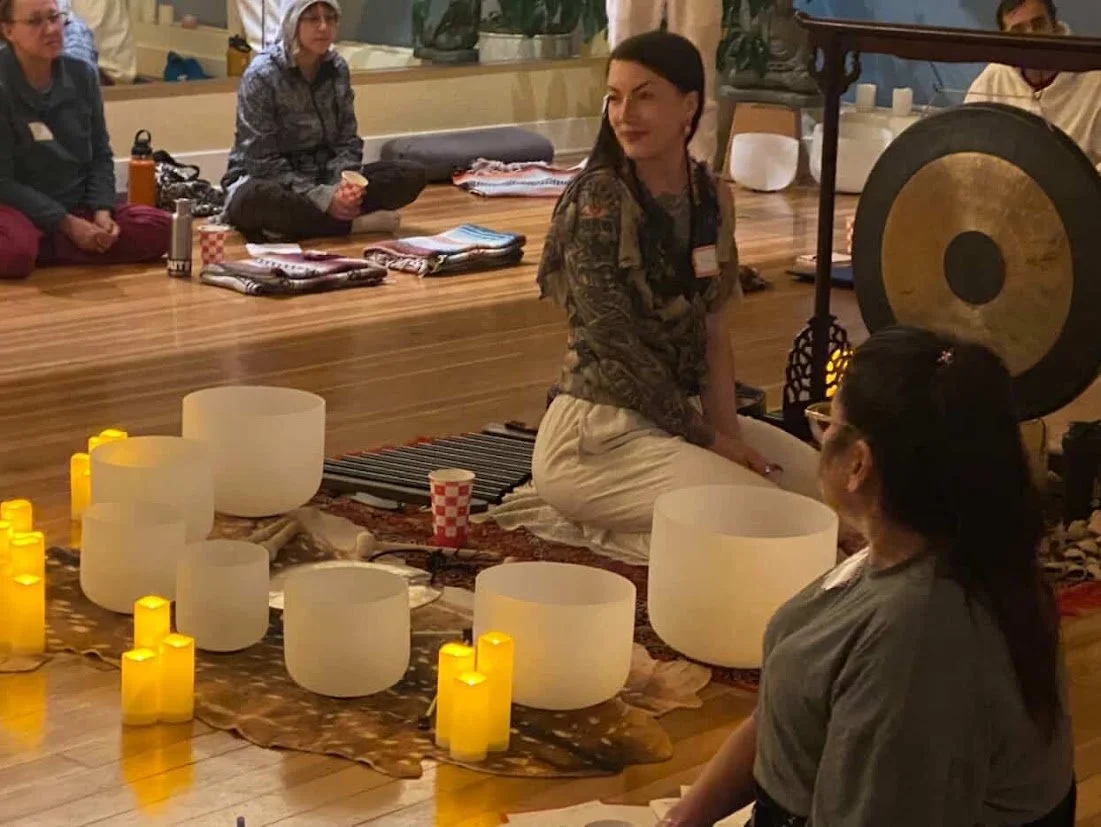 Women seated on a carpet with musical bowls and a gong, participating in a sound healing or meditation session, with candles around them.