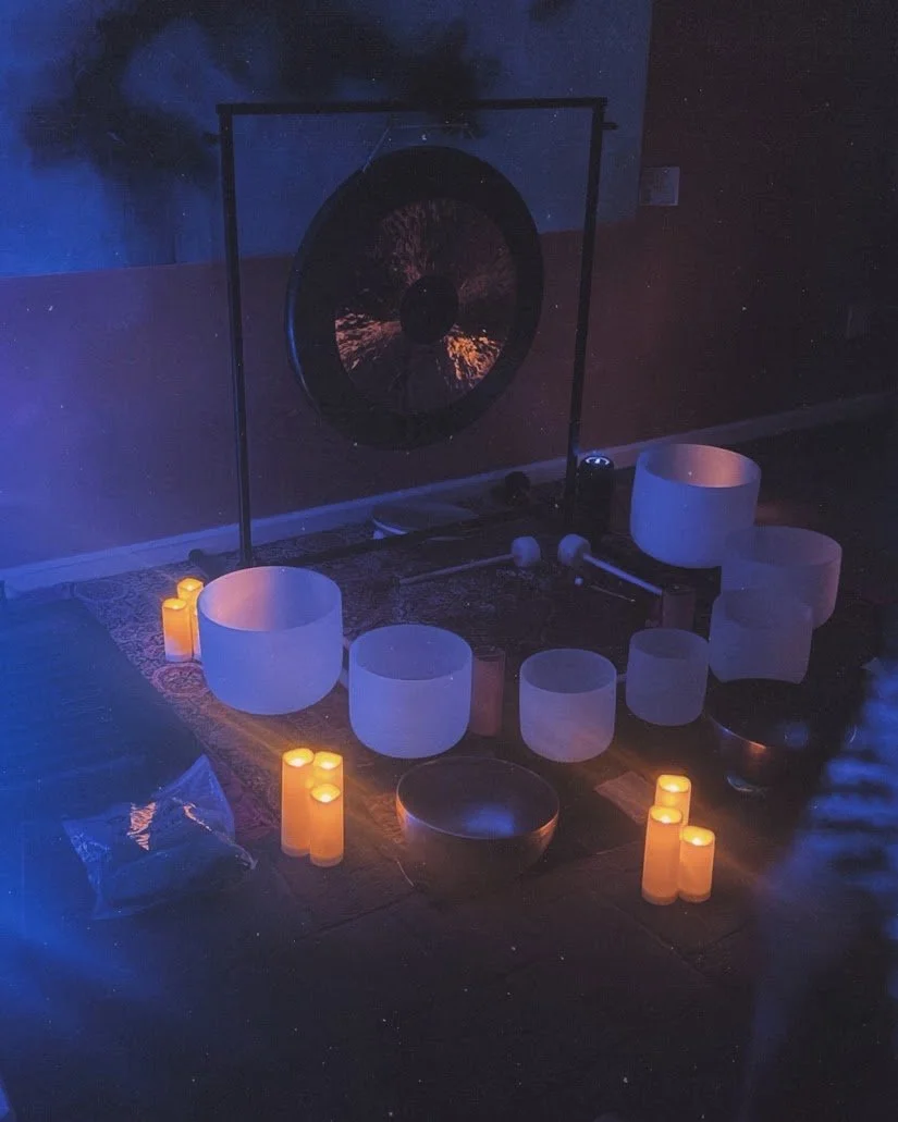 Dimly lit meditation or sound therapy room with crystal bowls, candles, and a gong.