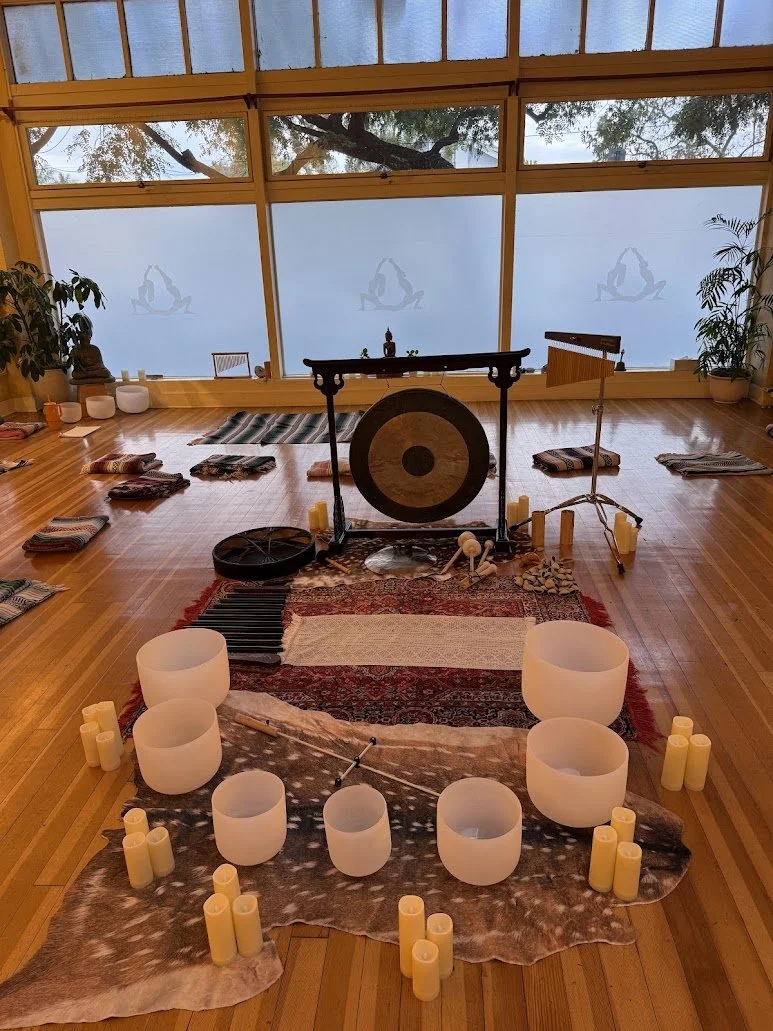 Interior of a yoga or meditation studio with bowls and candles arranged in a circle, a large gong in the center, wind chimes, and mats on the floor, with large windows showing trees outside.