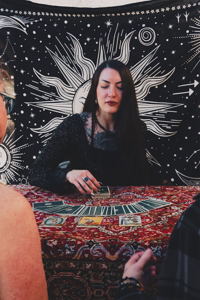 A woman with long dark hair and dark clothing sitting at a table with tarot cards, with a celestial-themed tapestry featuring a sun and stars behind her. Two other people are partially visible, one with a tan, forearm tattoo and the other with dark clothing.