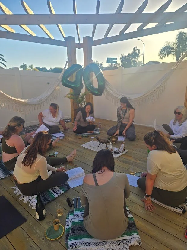 A group of women gathered outdoors, sitting on a wooden deck in a circle. There is a large silver balloon in the shape of the number 40, indicating a celebration. The background features a white fence, a hammock, and a pergola, with some trees and houses visible in the distance. The women are engaged in a discussion or activity, with papers and drinks around them.