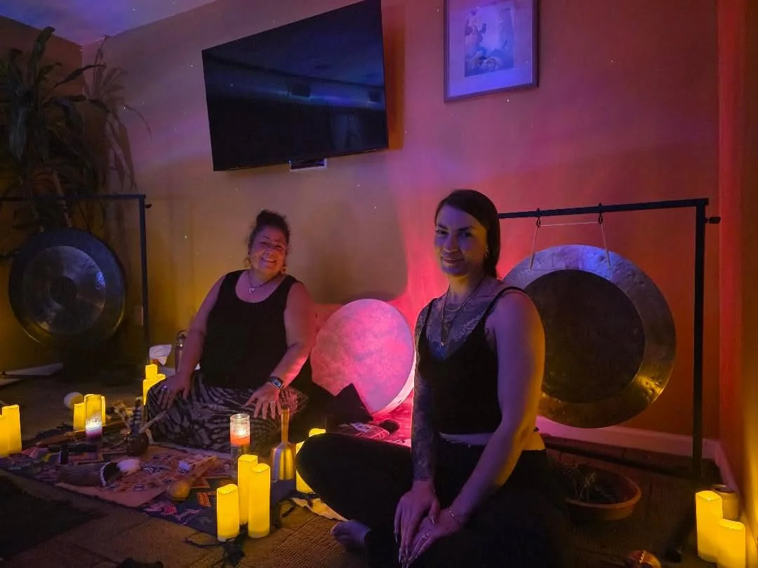 Two women sitting on the floor in a dimly lit room decorated with candles, gongs, and a large illuminated moon lamp, with a wall-mounted TV and picture frame in the background.