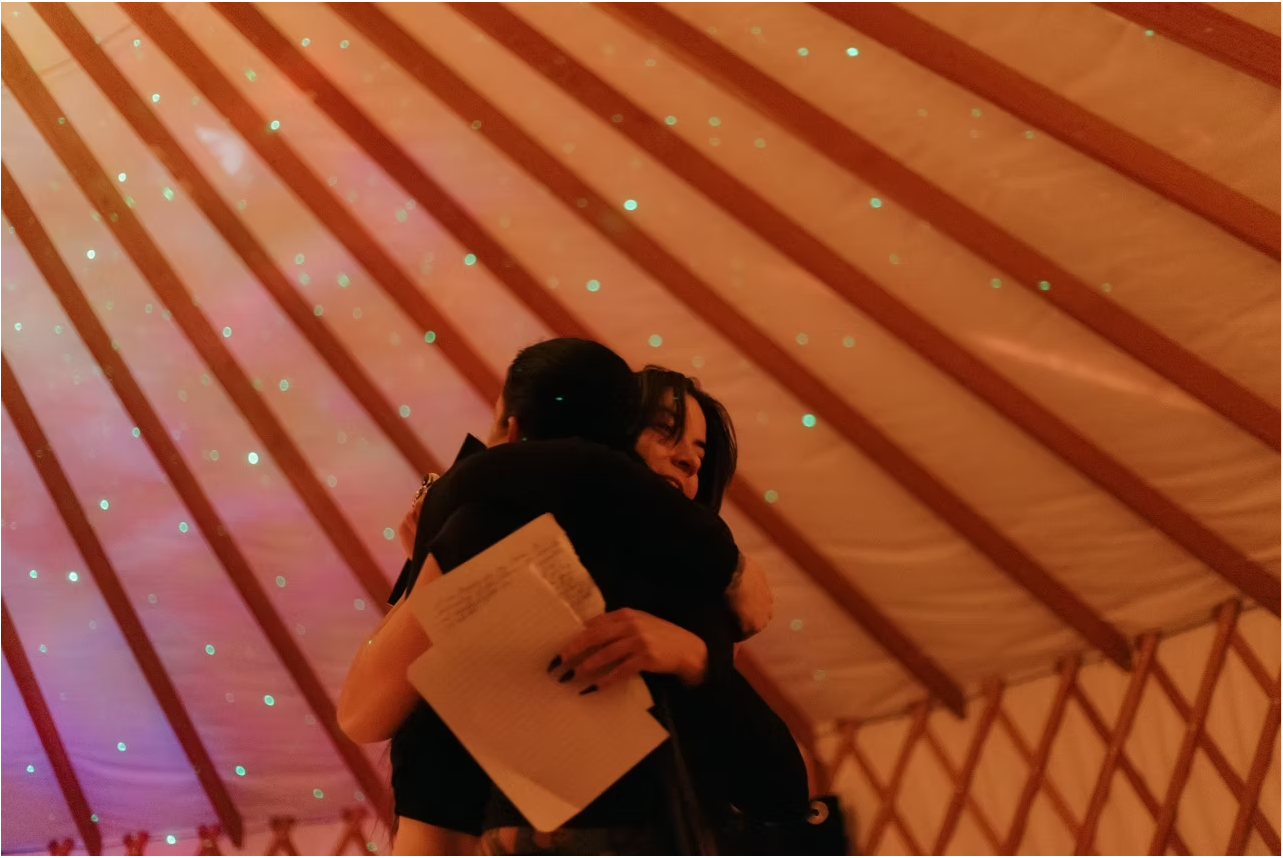 Two people hugging, one holding a piece of paper, inside a wooden ceiling structure with star-like lights.