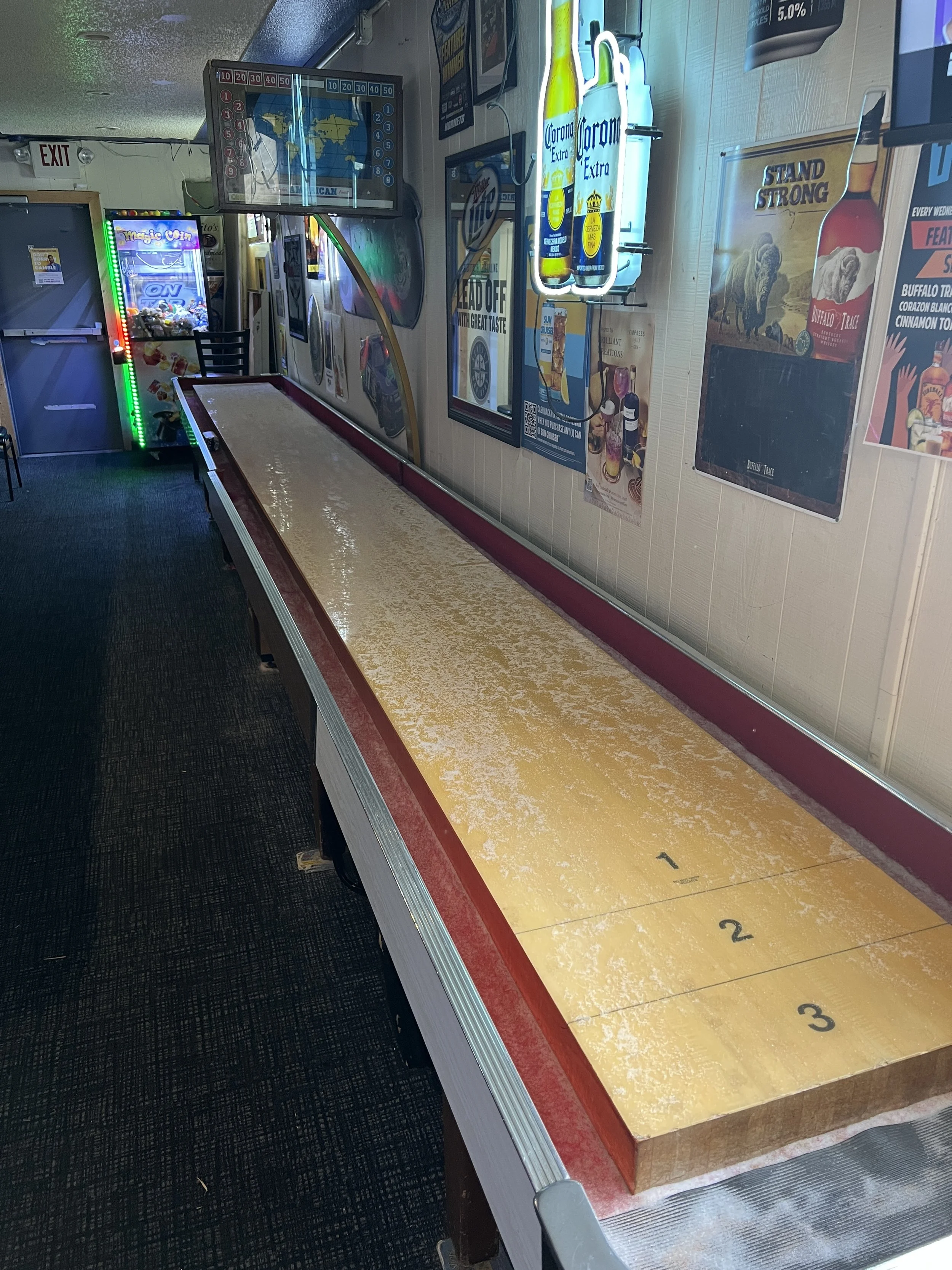 A shuffleboard game table in a bar or arcade, with a scored surface numbered 1 to 3, flanked by sports and beverage posters on the wall. An arcade game is visible in the background.