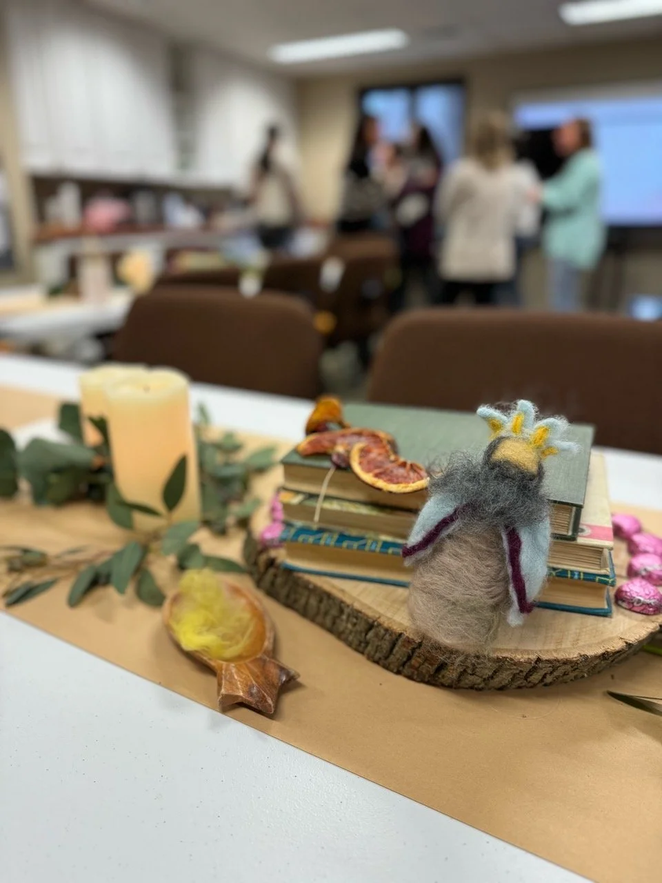 Decorative table setting for Thanksgiving with a candle, dried leaves, cotton candy, a small woolen figurine, and a stack of books on a wooden slab, with a blurred background of people in a room.