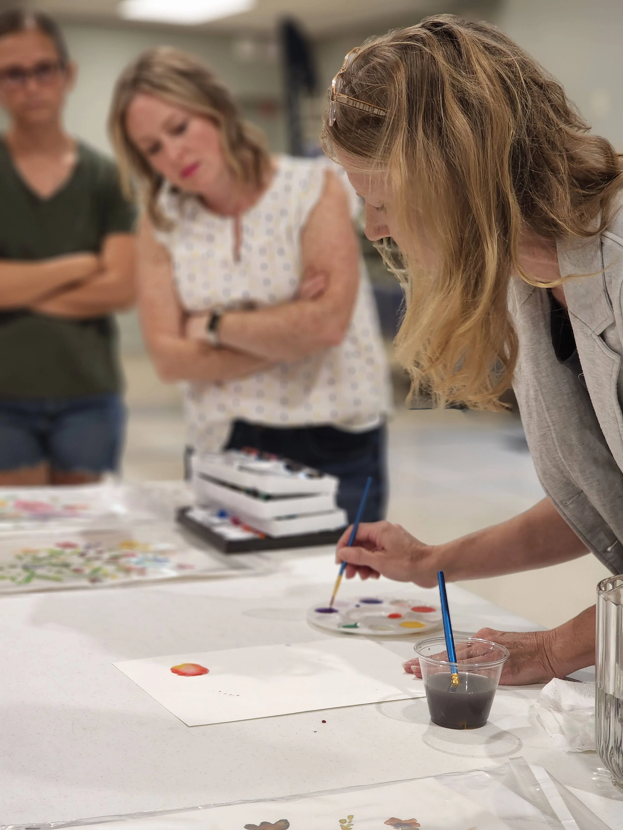 Woman painting with watercolors on paper at a table, while two spectators watch, in an indoor art workshop setting.
