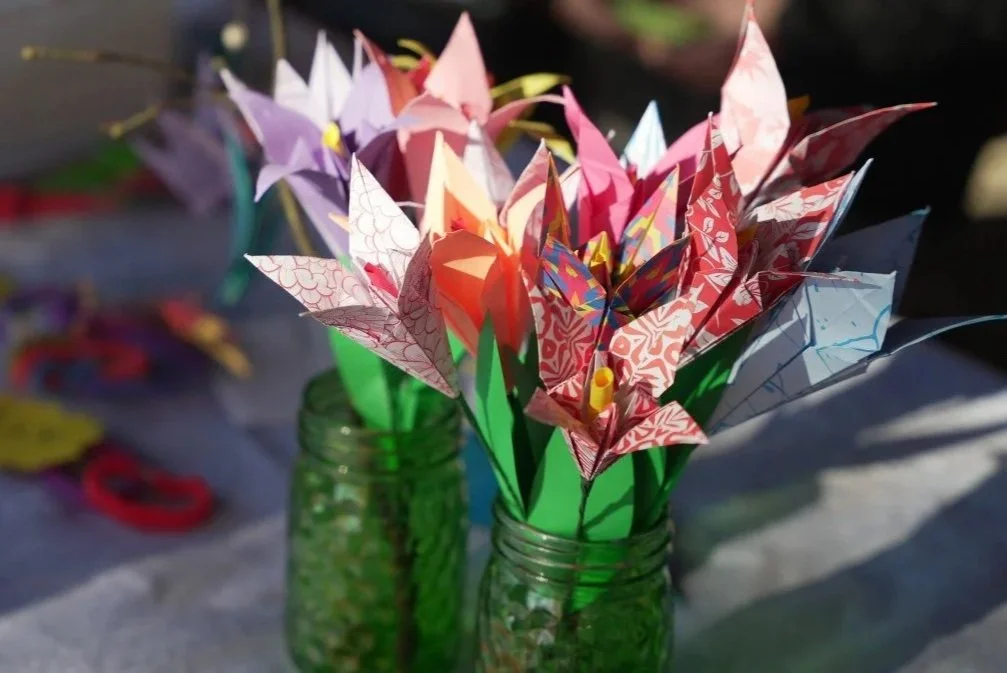 Colorful paper origami flowers in white, pink, red, purple, and blue, arranged in green vases on a table.