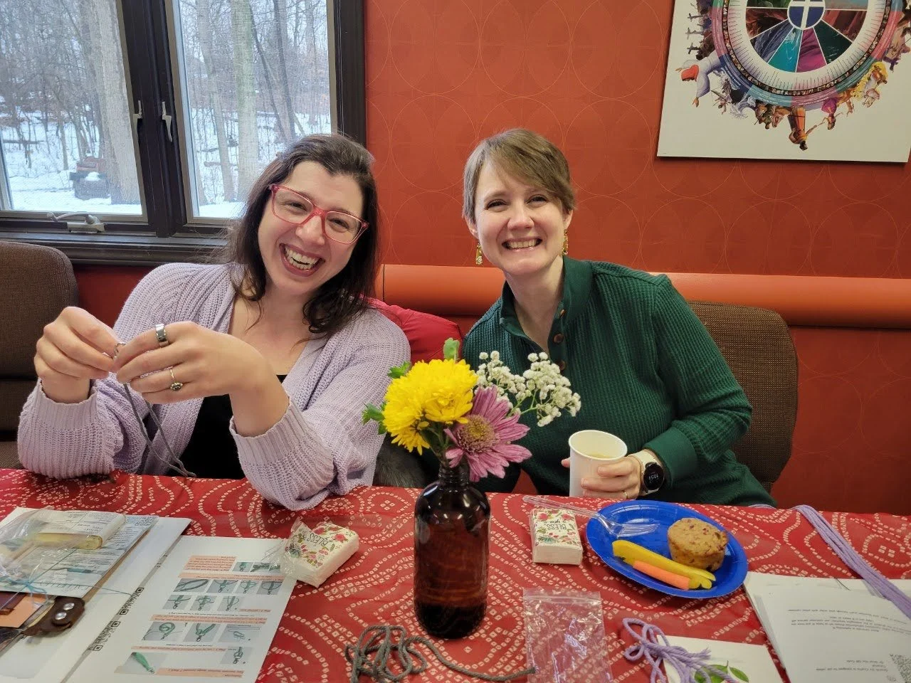 Two women sitting at a table with a vase of flowers, smiling, indoors with a window showing a snowy outdoor scene.