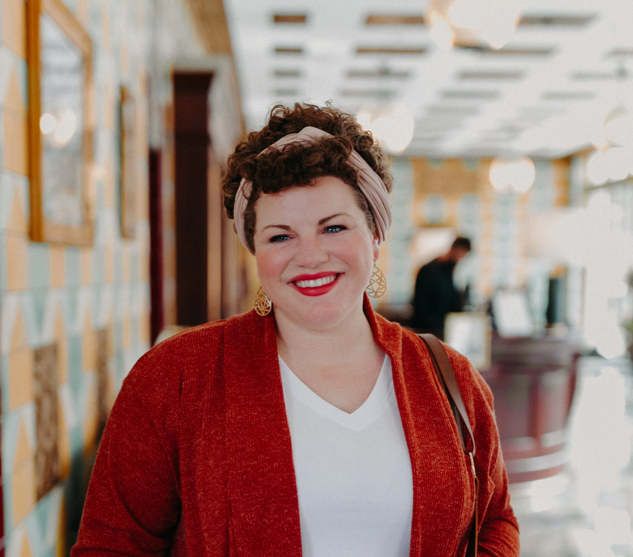 A woman with short curly hair, wearing a headband, red lipstick, a white V-neck shirt, a red jacket, and gold earrings, smiling inside a brightly lit cafe or restaurant.