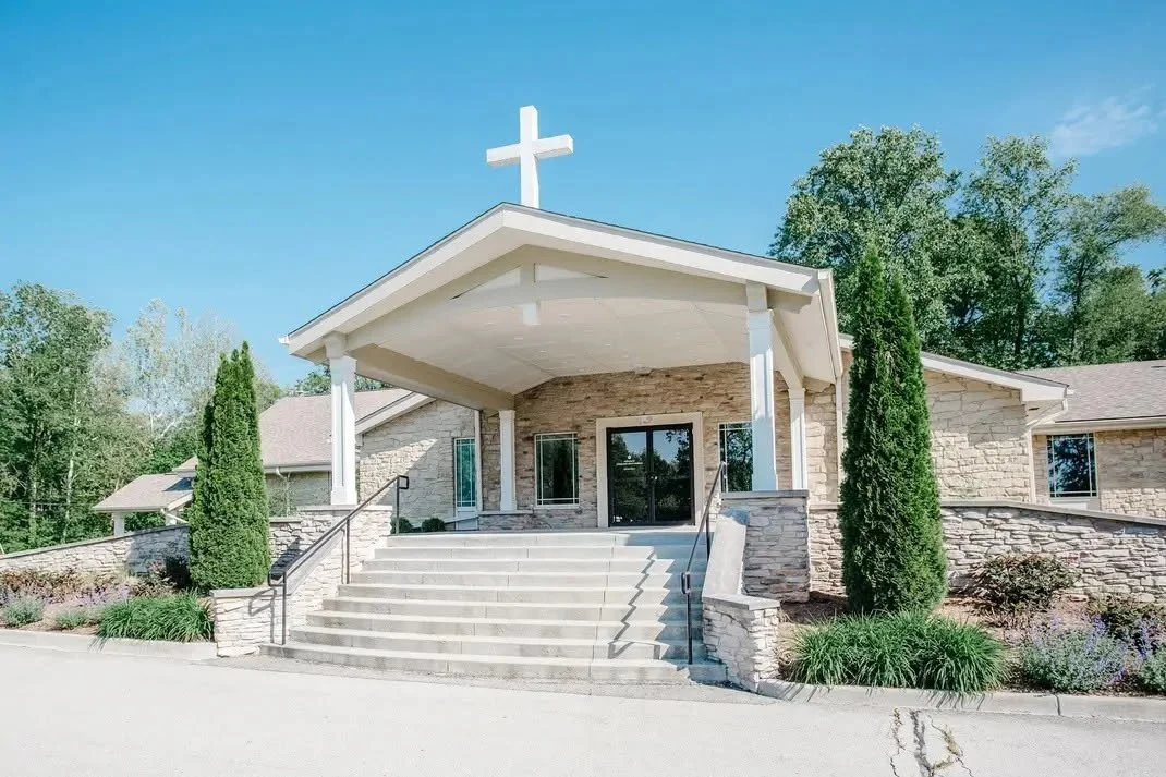 Front view of a church with a white cross on the roof, stone steps leading to the entrance, and lush greenery on either side.