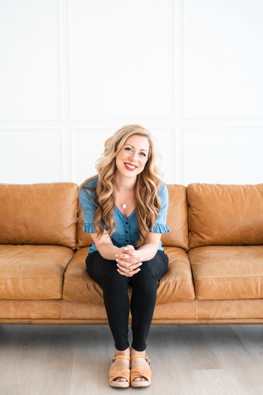 A woman with long, wavy blonde hair sitting on a brown leather sofa, smiling, wearing a blue button-up shirt, black pants, and tan sandals, in a bright room with white walls.