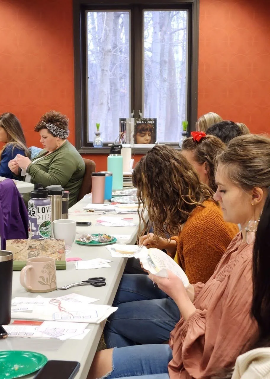 Women sitting at a long table in a room with an orange wall and a large window, engaged in a craft or sewing project.