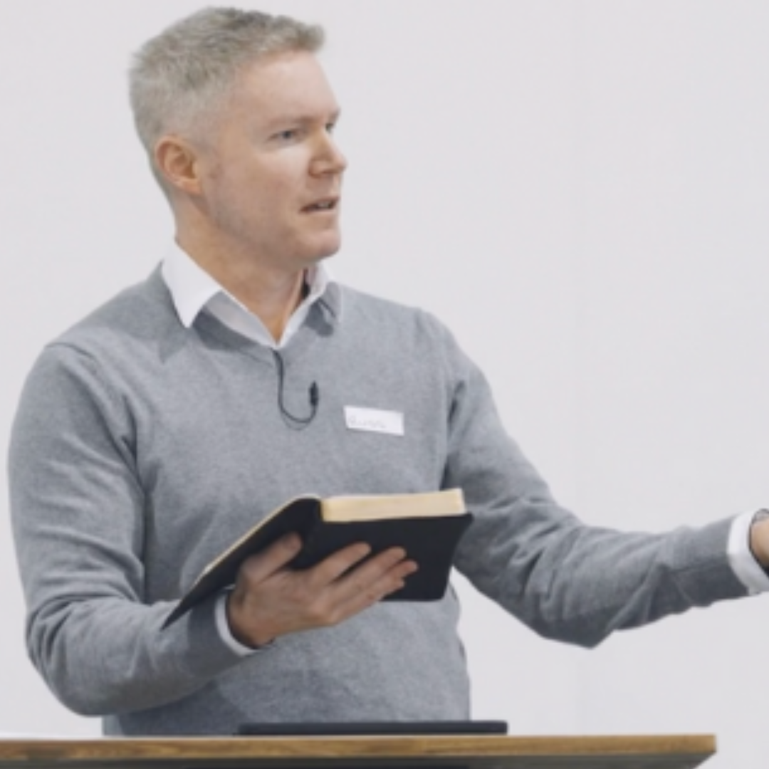 A man with light gray hair wearing a gray sweater over a white collared shirt, holding a notebook and speaking in front of a white wall.