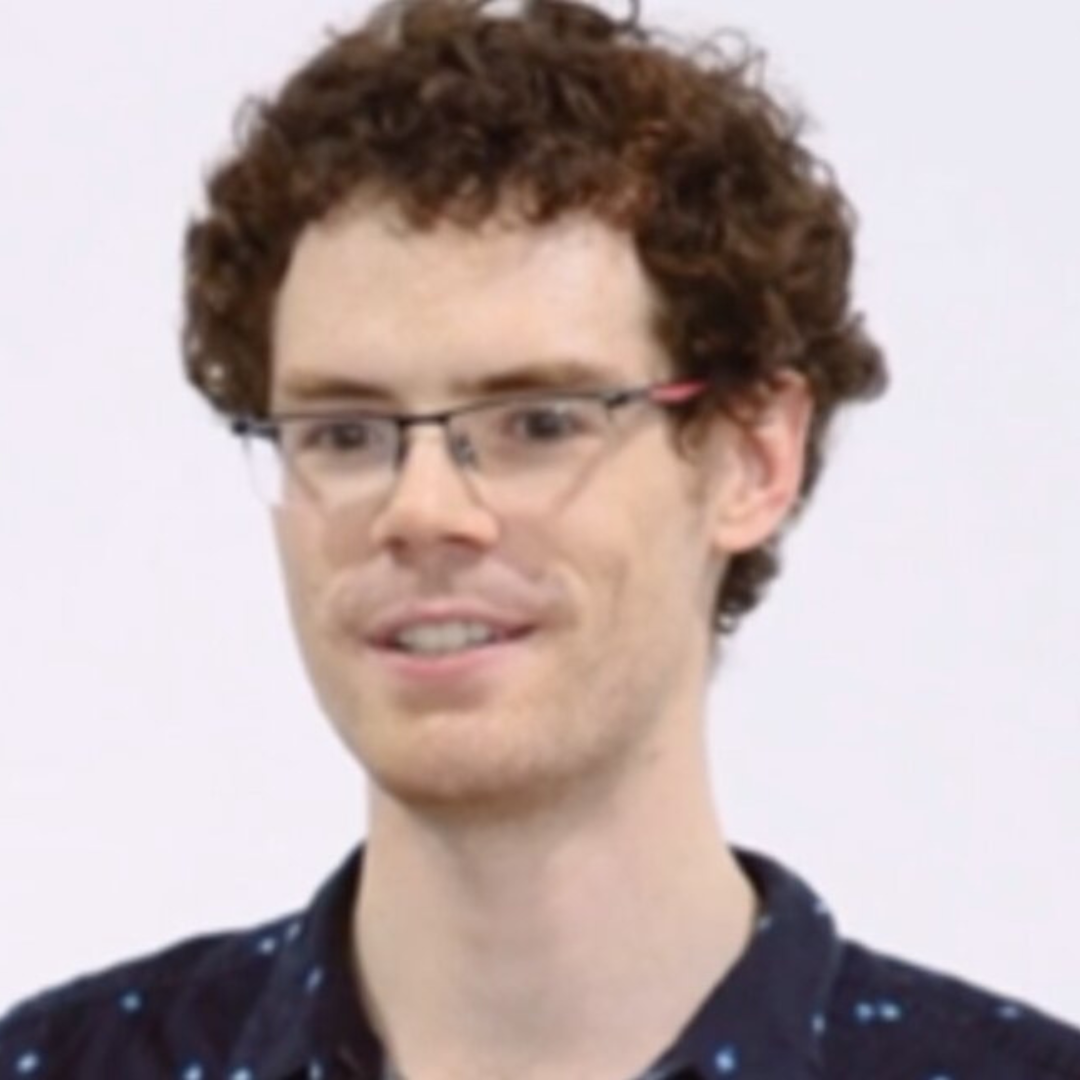 A young man with curly brown hair and glasses, wearing a dark shirt with small light-colored patterns.