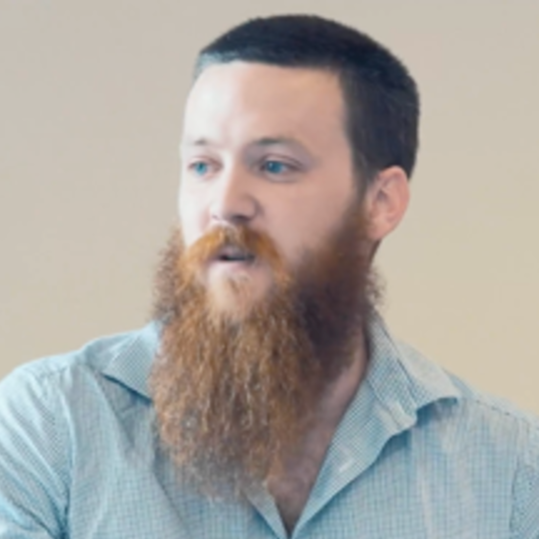 A man with a well-trimmed black hairstyle, bright blue eyes, and a full red beard, wearing a light gray checkered shirt, looking to the side against a plain beige background.