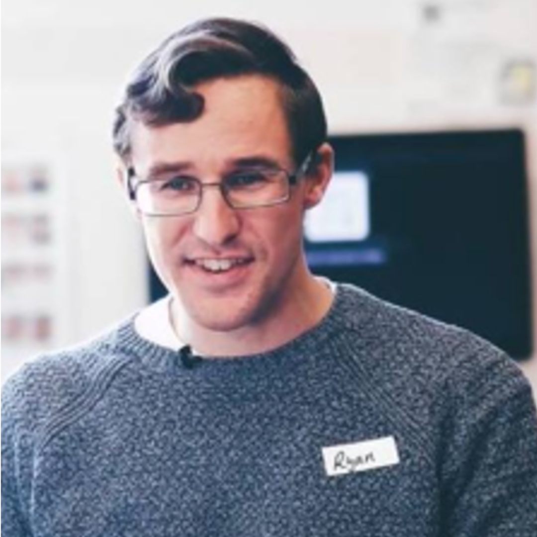 Young man with glasses smiling, wearing a gray sweater and a name tag that reads 'Ryan', standing indoors with a blurred computer screen in the background.