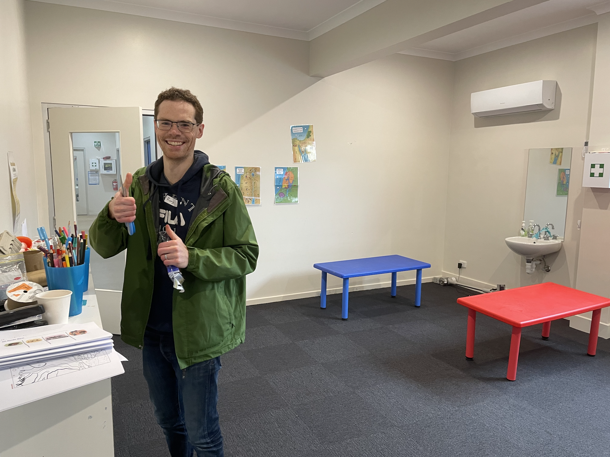 A smiling man in a green jacket and glasses giving a thumbs up in a room with colorful tables and children's artwork on the wall.