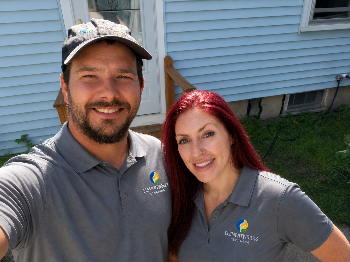 A man and woman taking a selfie outside a house with light blue siding, both wearing gray shirts with ElementWorks Cleaning logo, smiling at the camera.