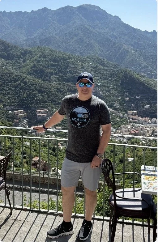 Man standing on a balcony with scenic mountain and town view behind him, wearing a cap, sunglasses, t-shirt, shorts, and sneakers.