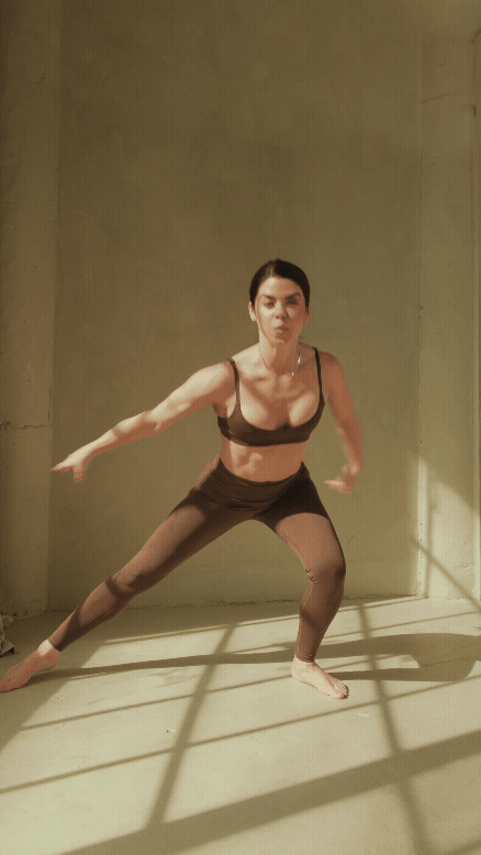 Woman in black athletic wear doing a wide-legged squat in a minimalistic room with sunlight and shadows.