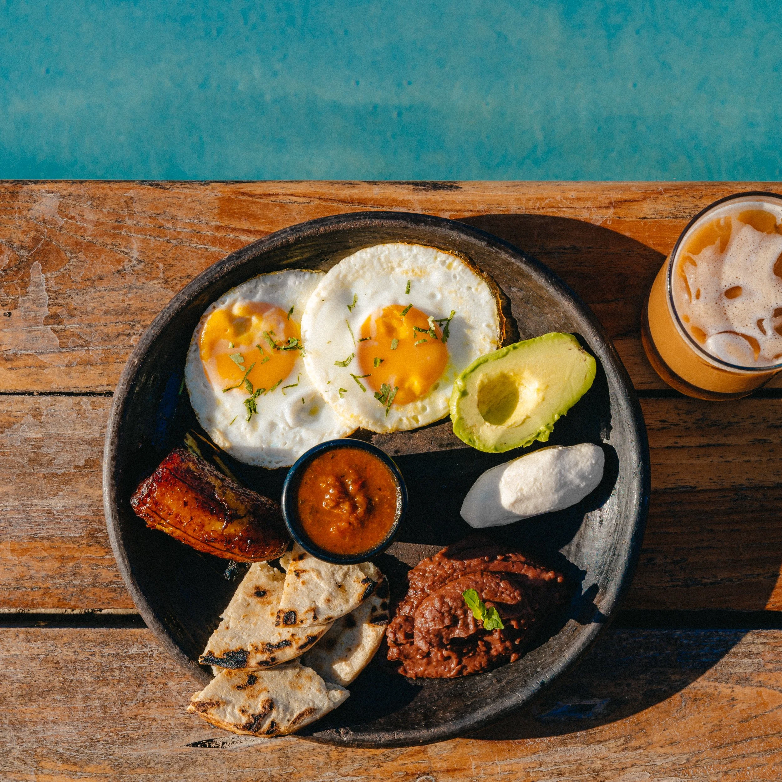 A round black plate with fried eggs, sliced avocado, grilled bread, and small bowls of salsa and chorizo on a rustic wooden table, with a blue background.