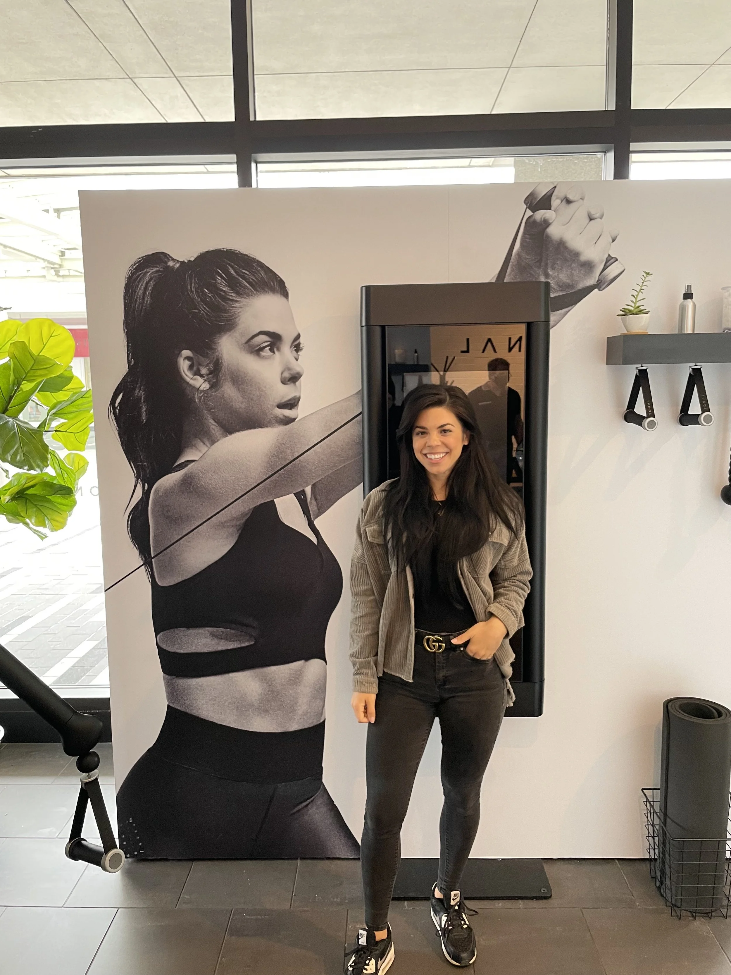 A smiling woman with long dark hair standing in front of a large black and white poster of a female athlete holding a bat, inside a modern gym or fitness center.