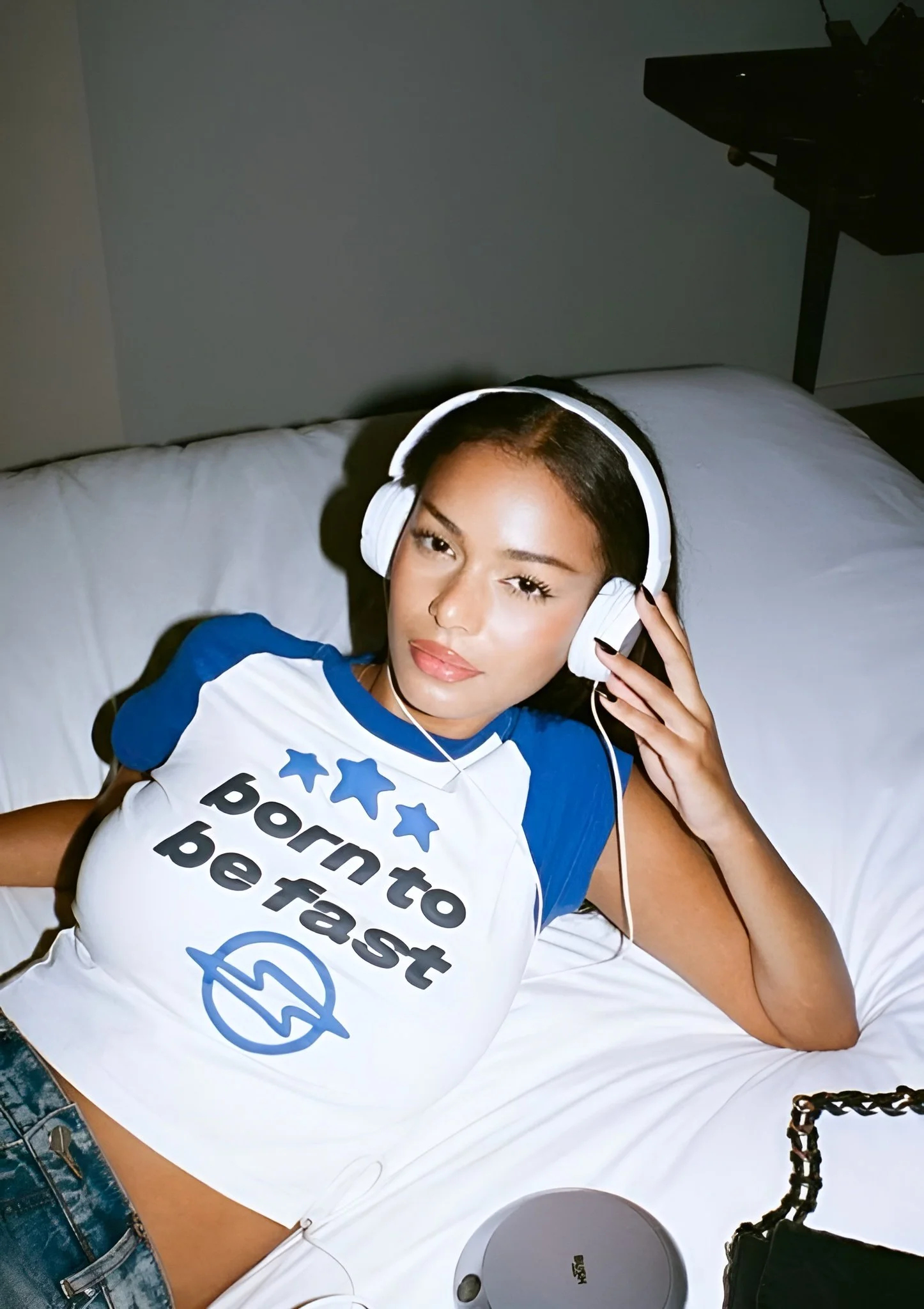 A young woman with dark hair and light brown skin, lying on a white bed, wearing a blue and white t-shirt that says 'born to be fast' with blue stars and a lightning bolt symbol, listening to music with large white headphones, looking at the camera.