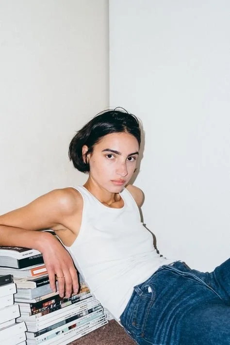 Young woman with short dark hair wearing a white tank top and jeans sitting on the floor against a white wall, leaning on a stack of books.