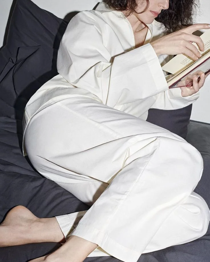 A woman with dark curly hair reading a book while lying on her side on a bed, wearing white pajamas.
