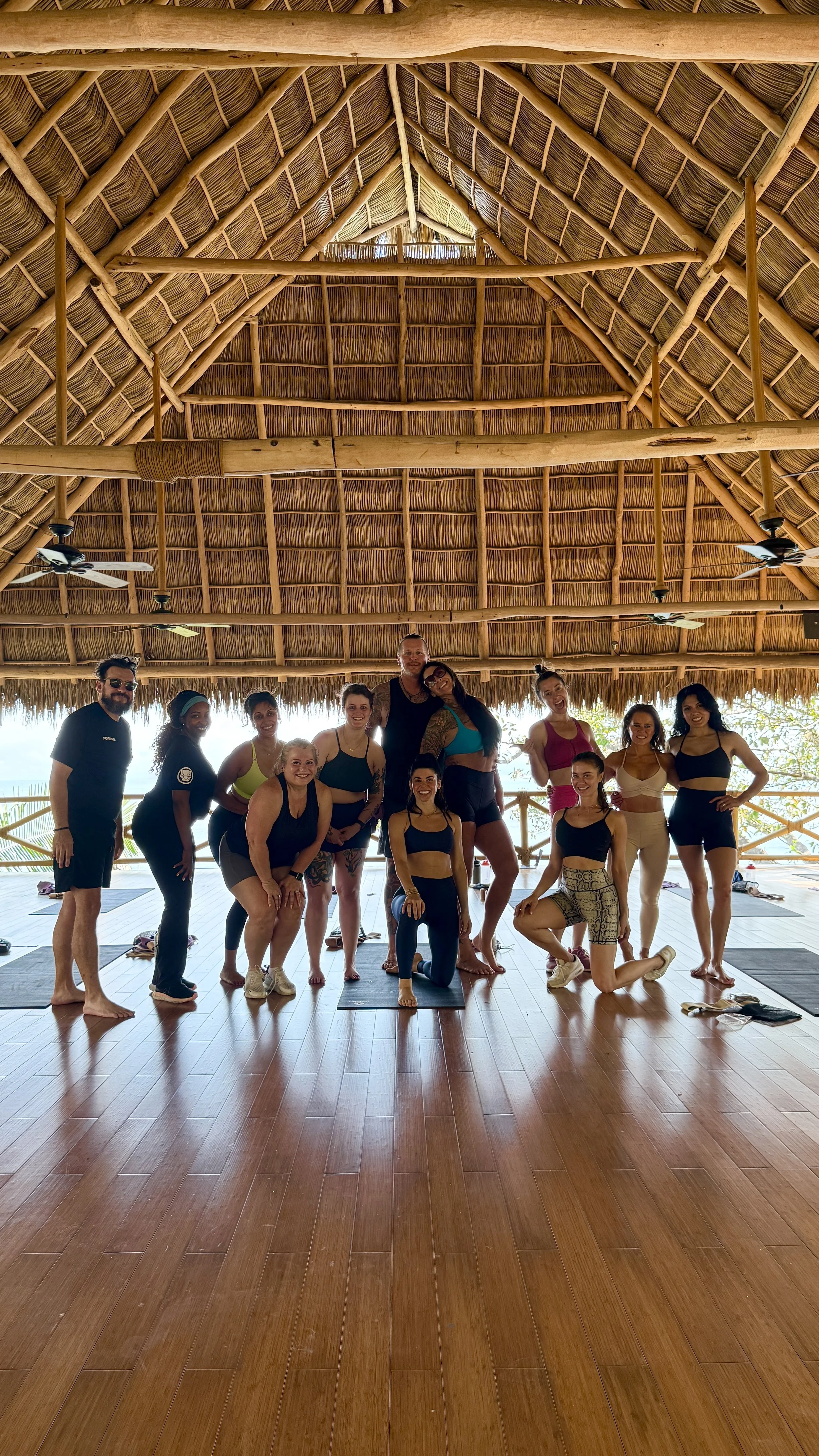 Group of people in yoga attire posing in a yoga studio with a thatched roof, wooden floor, and open sides with a view of nature outside.