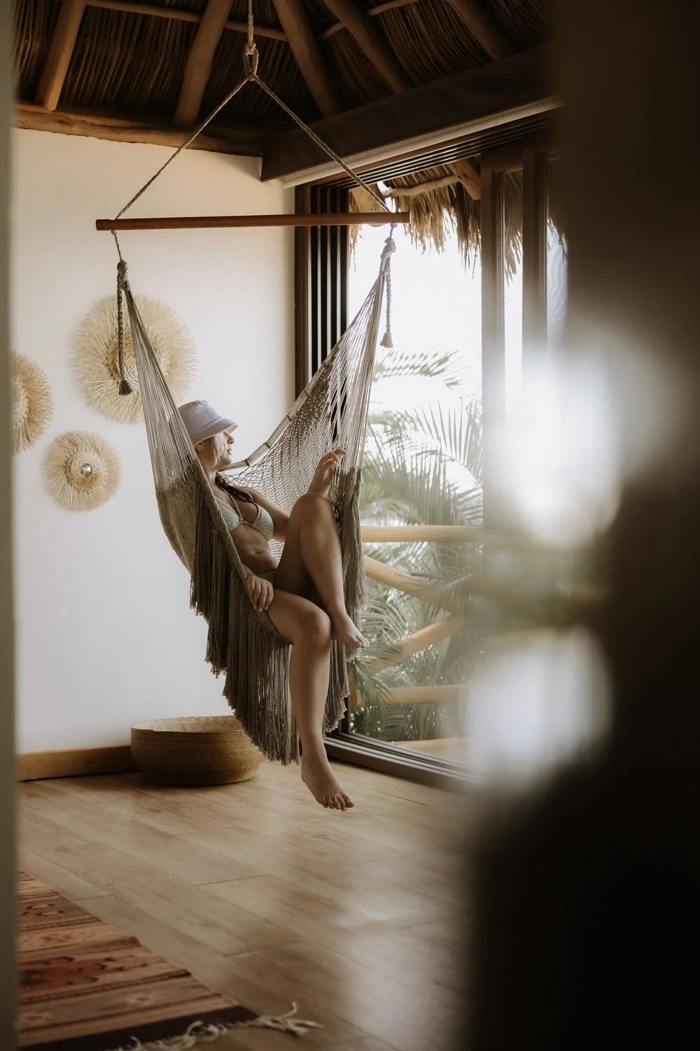 A woman relaxing on a hanging hammock chair indoors near a large window with a view of tropical plants outside.