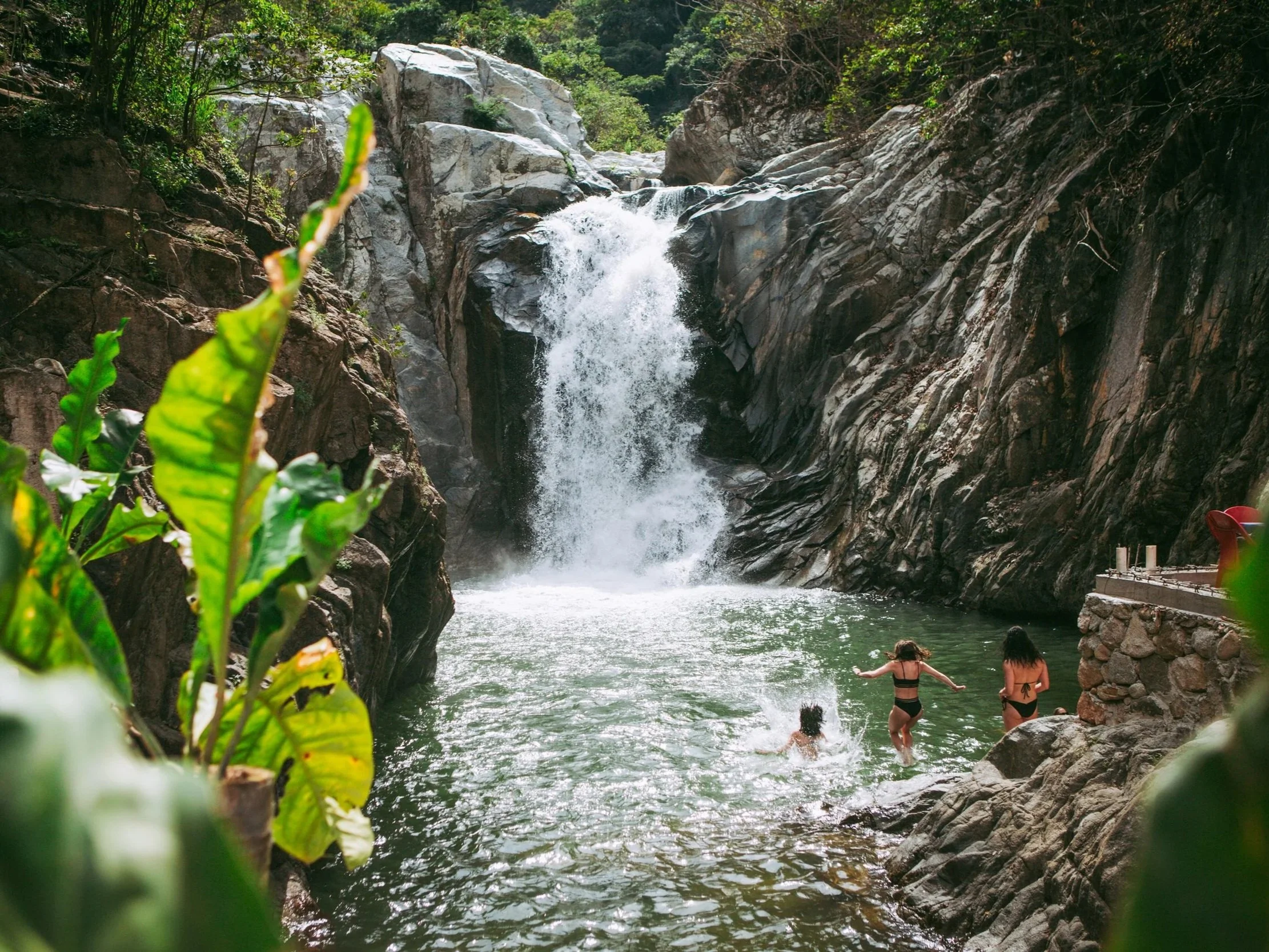 People swimming and relaxing in a natural waterfall pool surrounded by rocky cliffs and lush greenery.