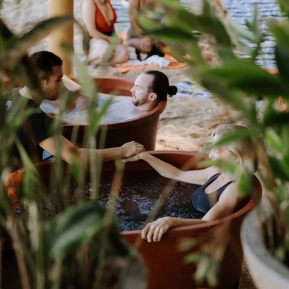 People relaxing in hot tubs outdoors, holding hands, with tropical plants in the foreground.