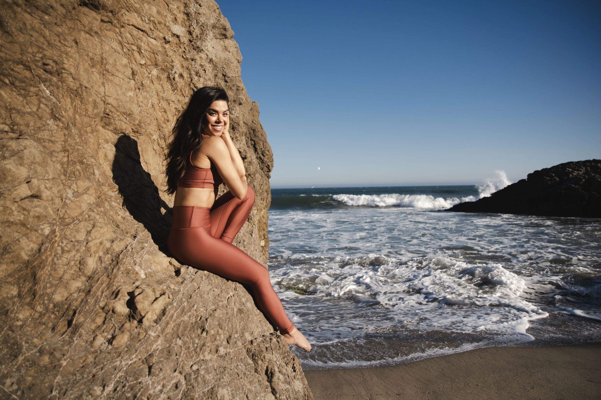 Woman in a rust-colored athletic outfit sitting on a large rock on a sandy beach with ocean waves and a clear blue sky in the background.