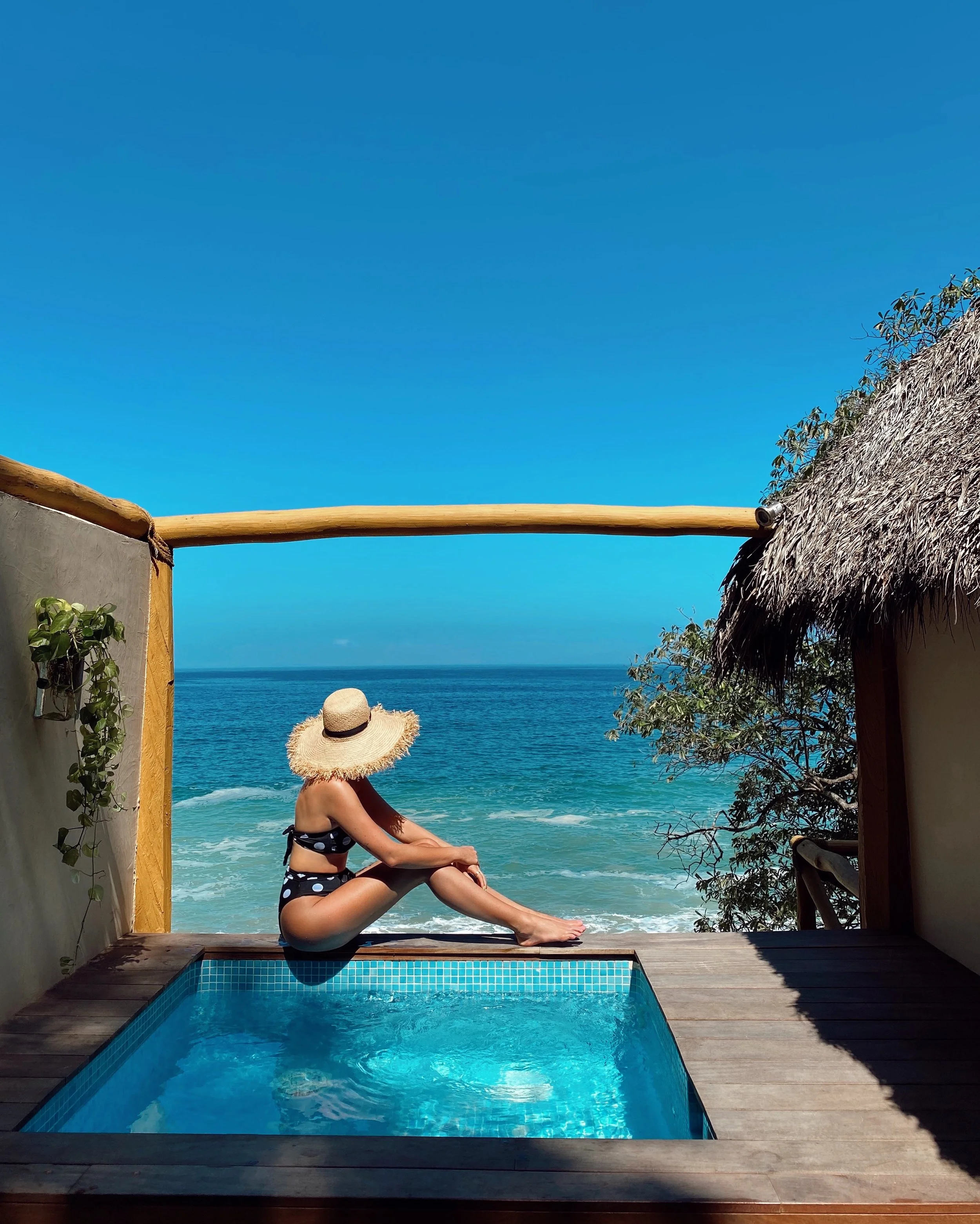 A woman in a black polka dot bikini and a wide-brimmed straw hat sitting by a small pool overlooking the ocean on a sunny day