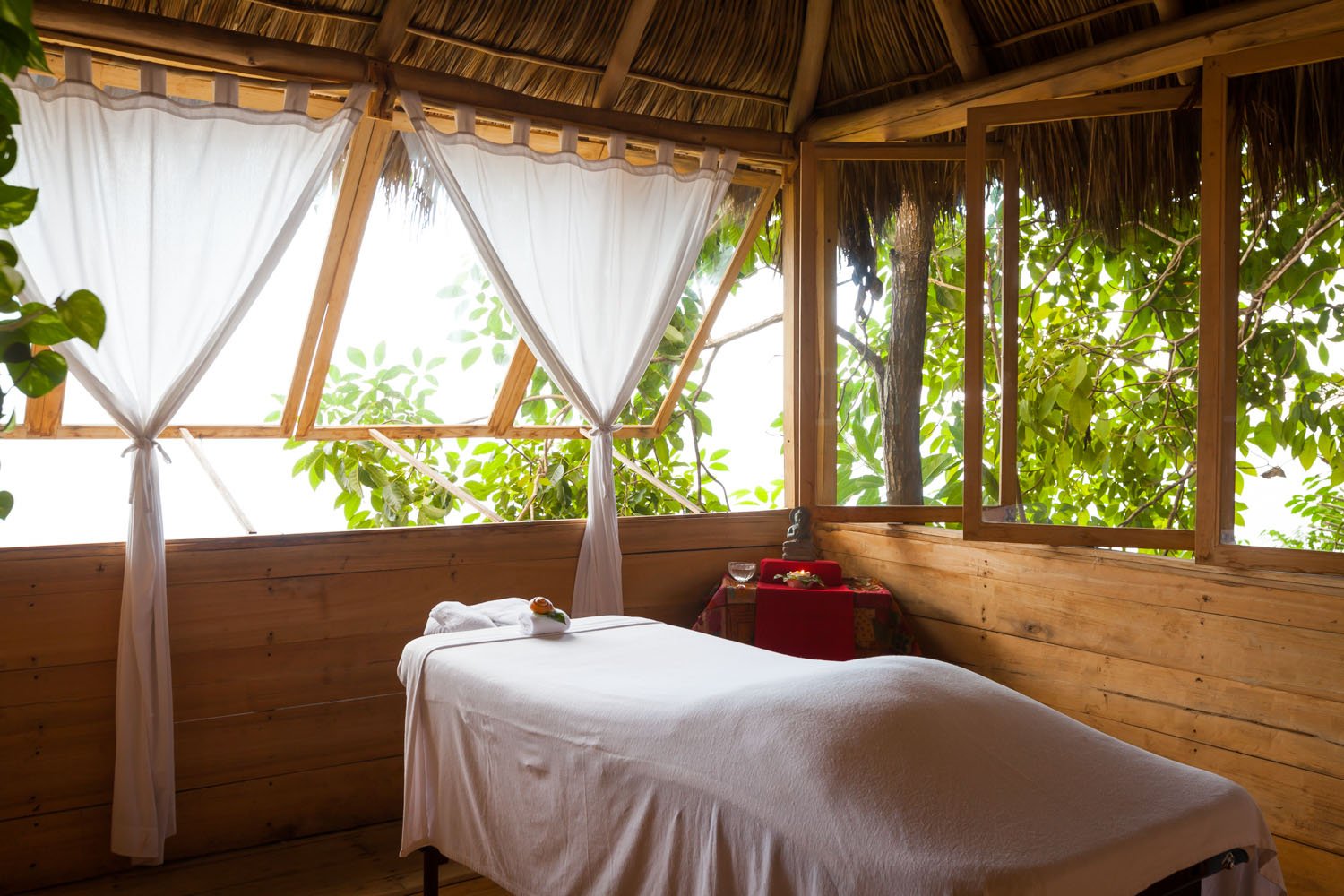 A massage table in a wooden room with large windows and open curtains, overlooking green foliage.