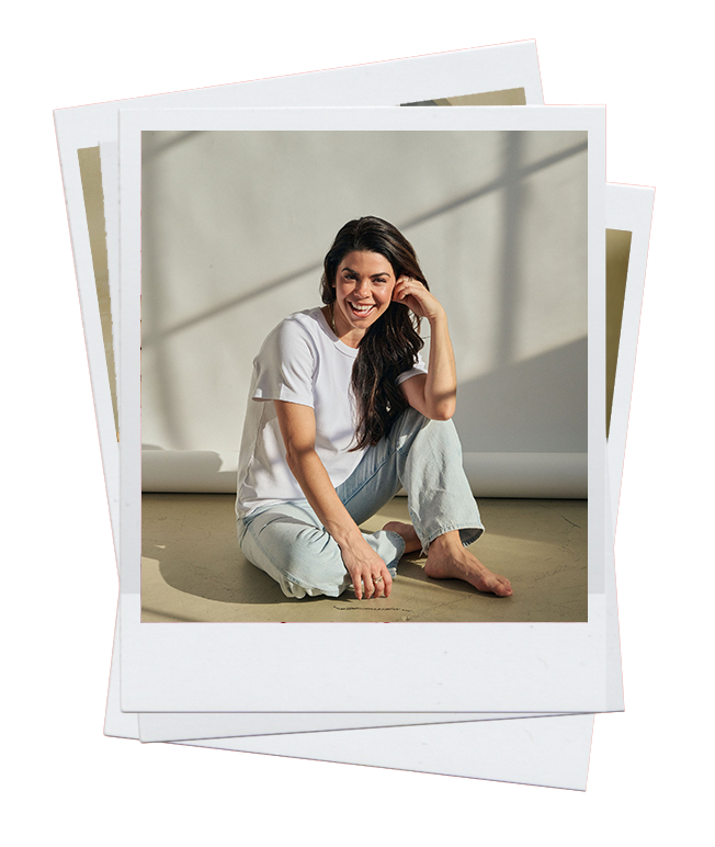 Woman sitting on the floor in a brightly lit room, smiling and wearing a white t-shirt and light jeans.